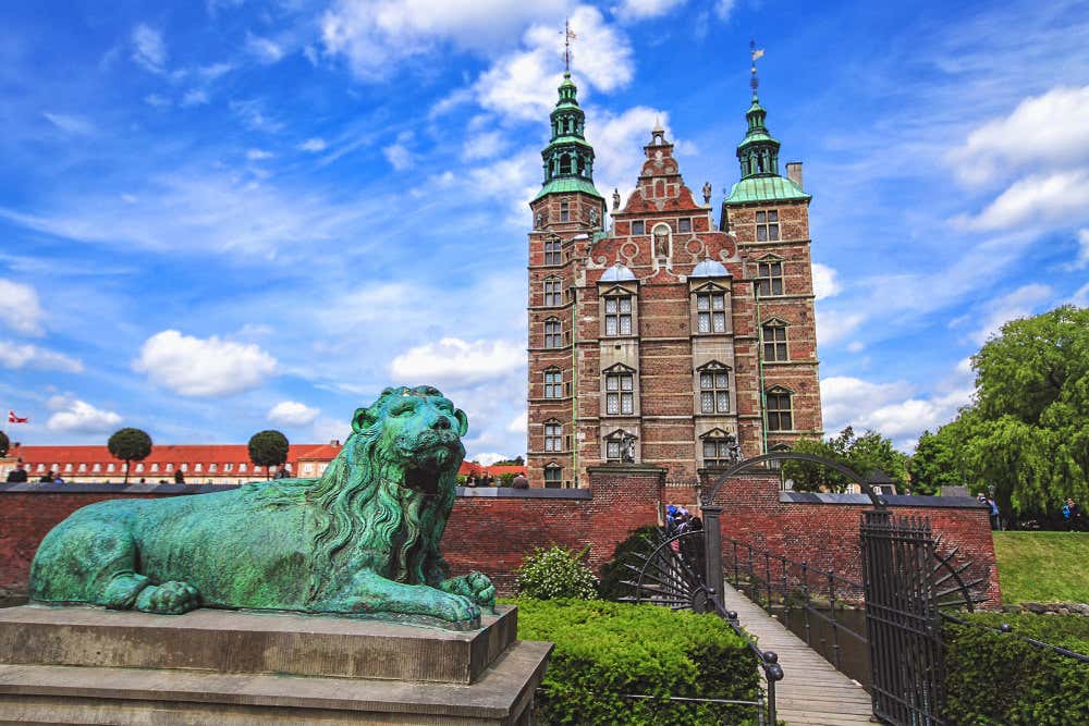 Castelo de Rosenborg, em Copenhague. Castelo alto, feito de tijolos laranjas com torres esverdeadas nas extremidades. No primeiro plano, na parte inferior esquerda, há uma estátua de um leão, também esverdeado, para representar Dinamarca.